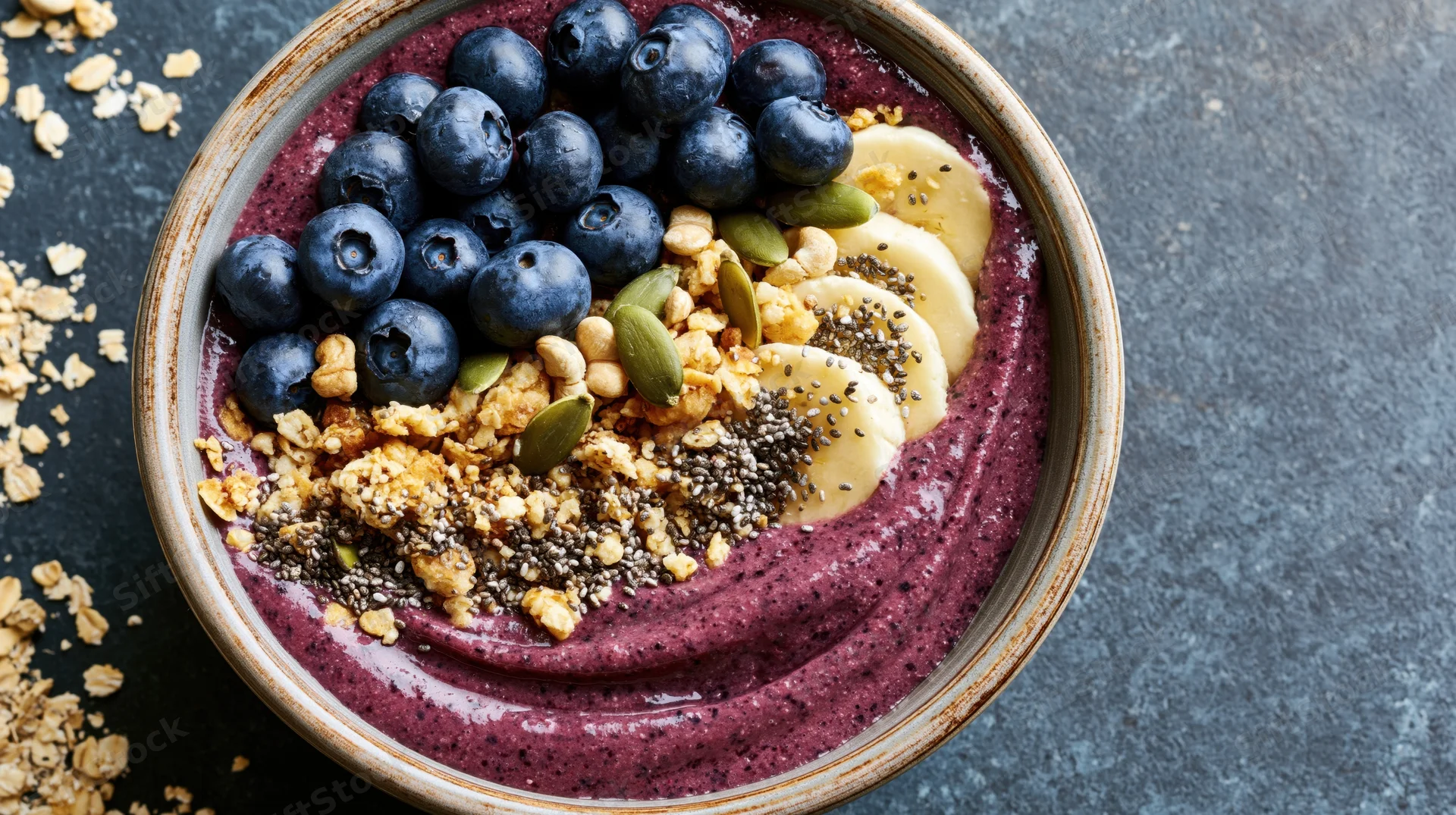 Acai smoothie bowl topped with blueberries, banana slices, granola, and seeds on a dark stone surface