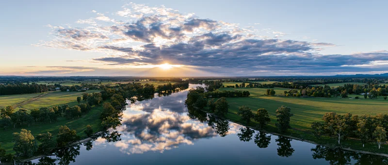 Aerial drone view of a river winding through green farmland at sunset with clouds reflected in the water