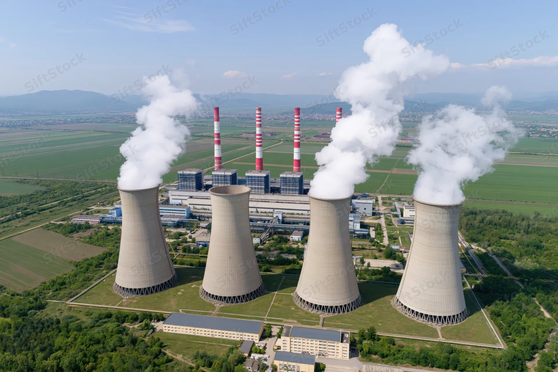Aerial view of a large coal-fired power plant with cooling towers emitting steam, surrounded by green fields and distant mountains
