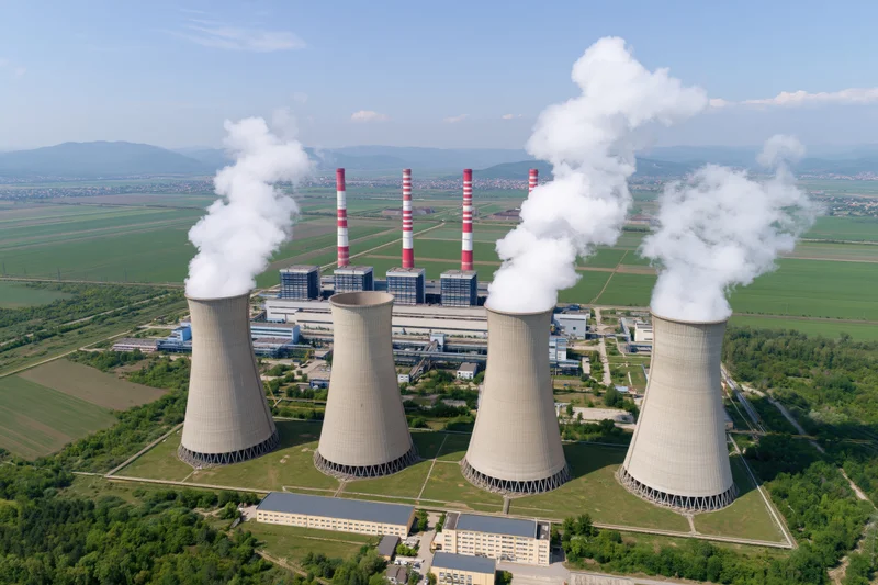 Aerial view of a large coal-fired power plant with cooling towers emitting steam, surrounded by green fields and distant mountains