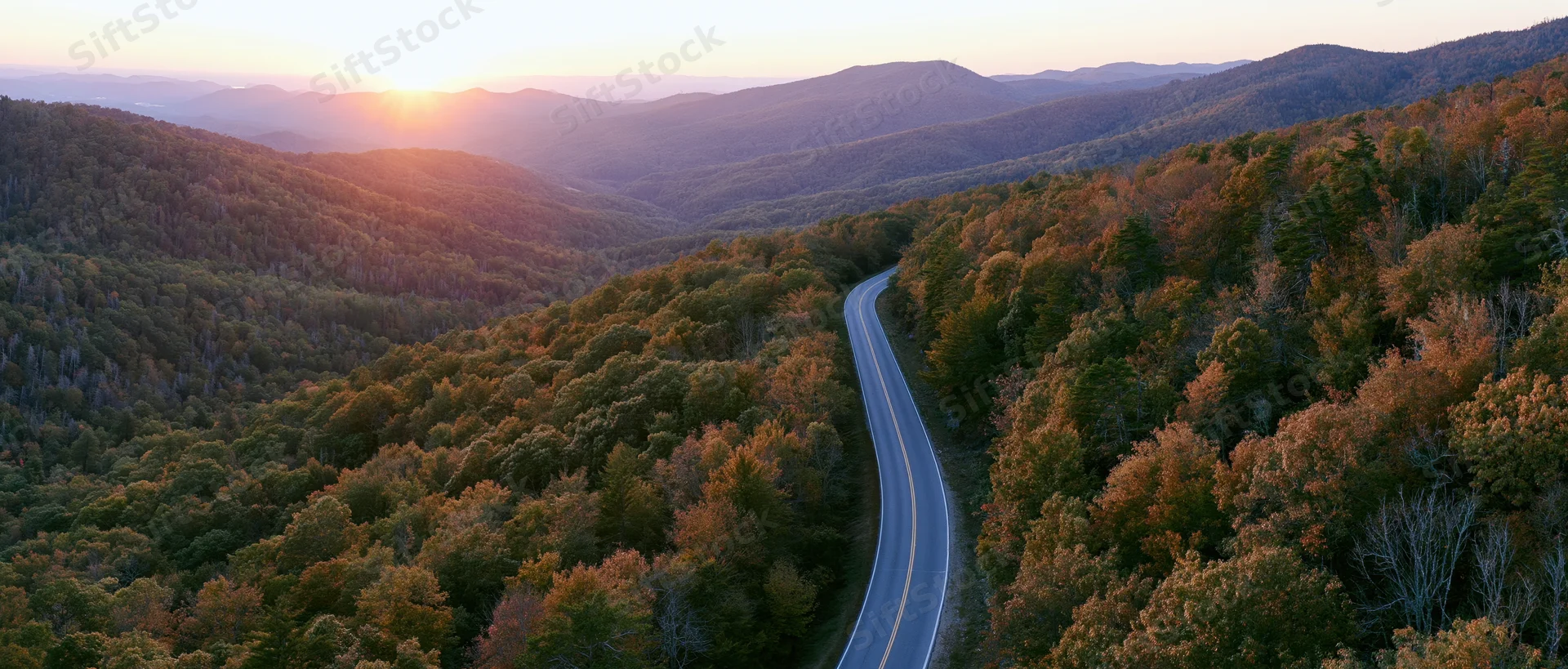 Aerial view of a winding two-lane road through autumn forested mountains at sunset