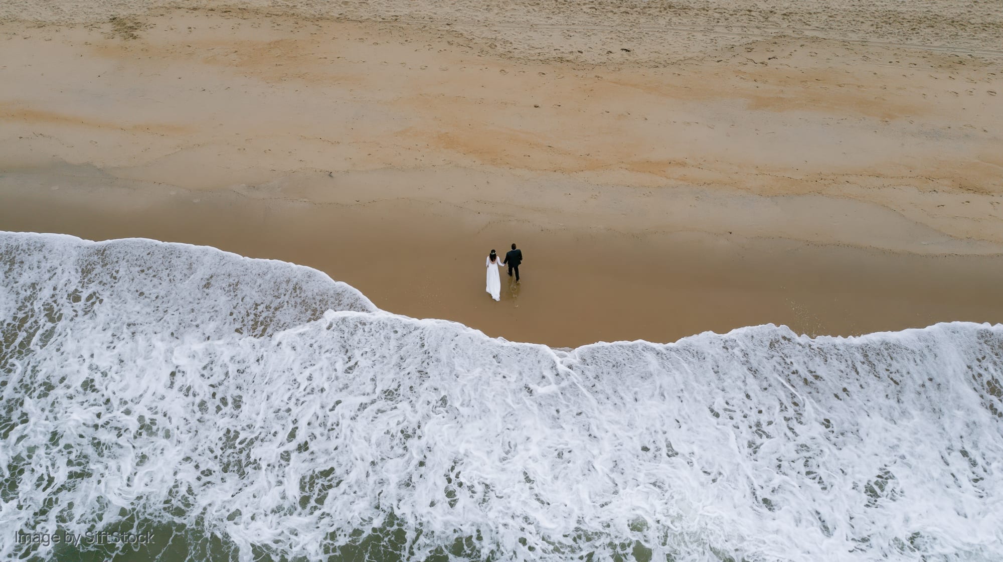 Bride and groom walking along the beach near crashing waves, aerial view