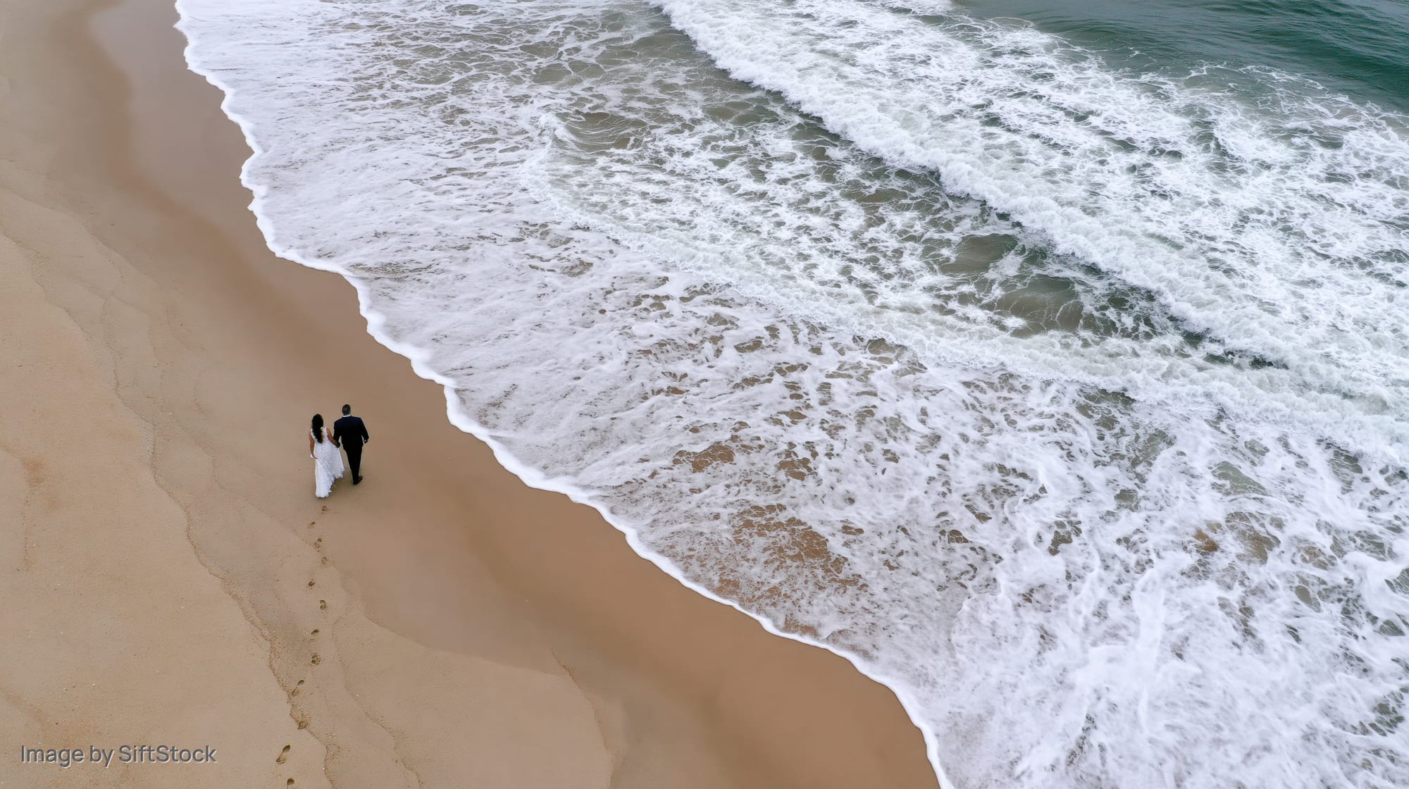 Couple walking along sandy beach beside crashing waves, aerial view
