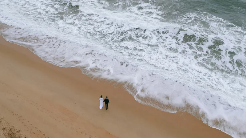 Couple walking along sandy beach near crashing waves from aerial view