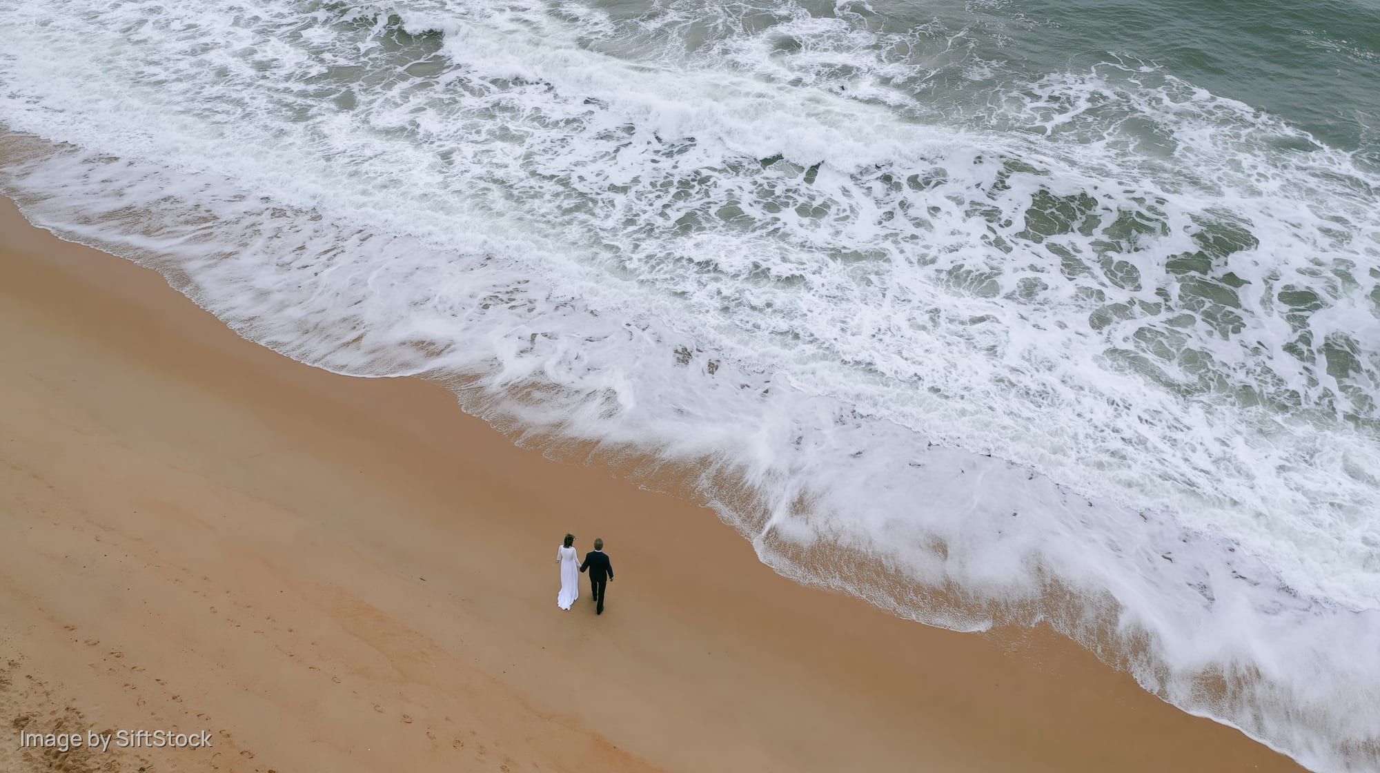 Couple walking along sandy beach near crashing waves from aerial view