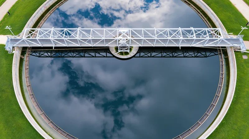 Aerial drone view of circular water reservoir with metal walkway bridge and sky reflection