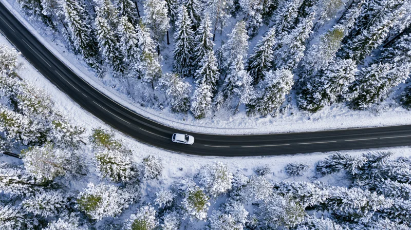 White car driving on curved road through snow-covered forest from aerial view