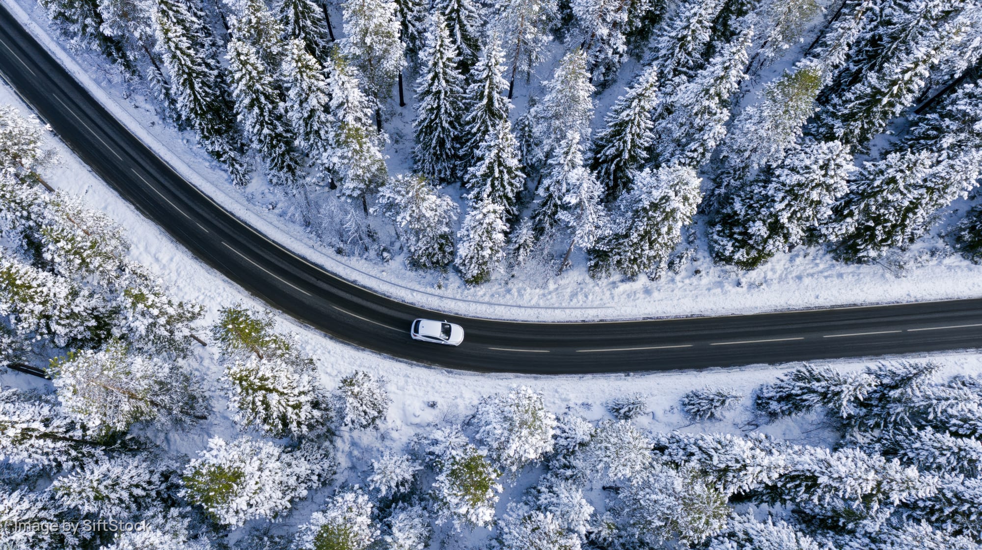 White car driving on curved road through snow-covered forest from aerial view
