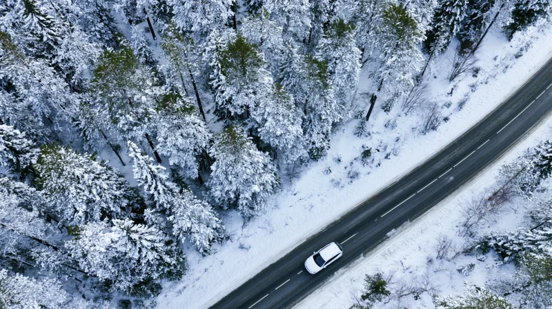 White car driving on a snowy road through a snow-covered forest from an aerial view