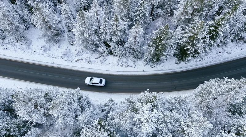 White car driving on a curved snow-covered road through a forest viewed from above