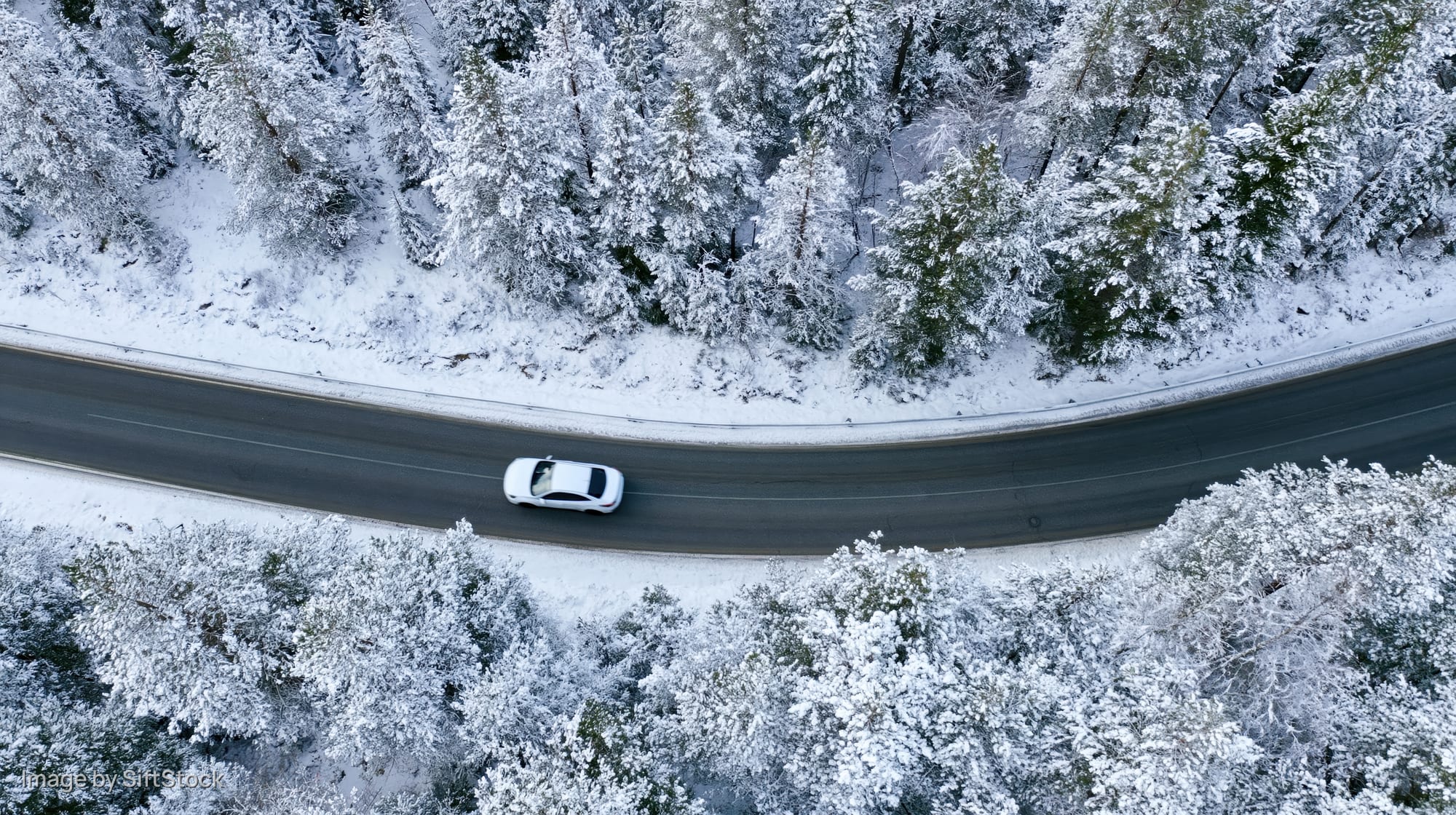 White car driving on a curved snow-covered road through a forest viewed from above