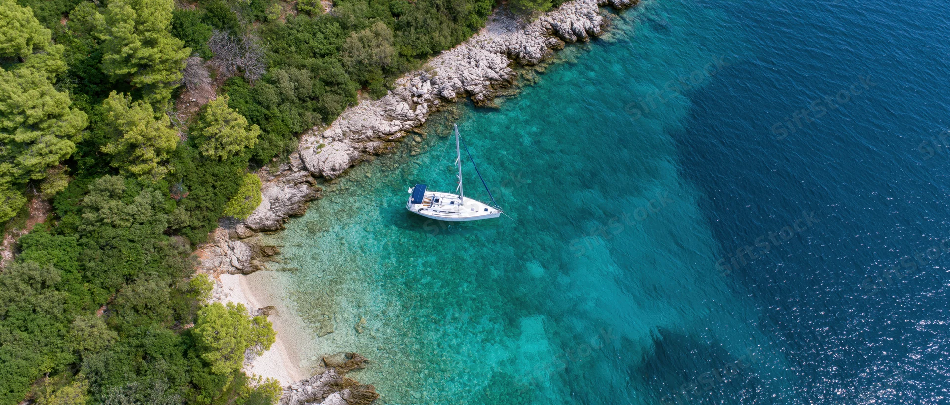 Aerial view of a white sailboat anchored in a turquoise cove beside a rocky shore with green trees and a small beach