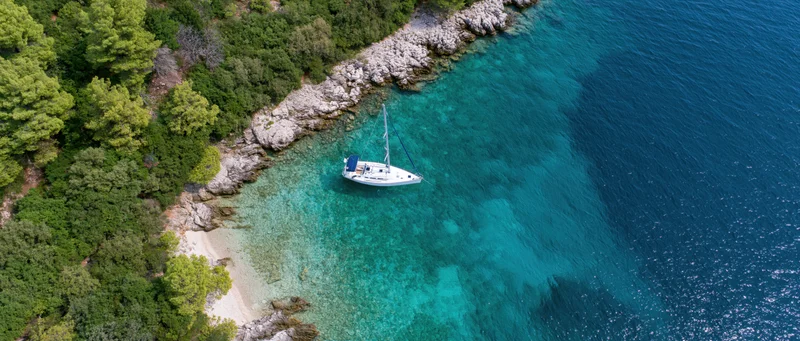 Aerial view of a white sailboat anchored in a turquoise cove beside a rocky shore with green trees and a small beach