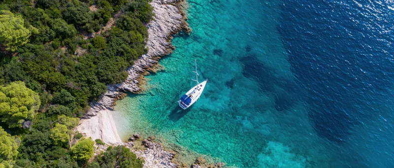 Aerial drone view of a sailboat anchored in a secluded cove with turquoise water beside a rocky shore and green trees