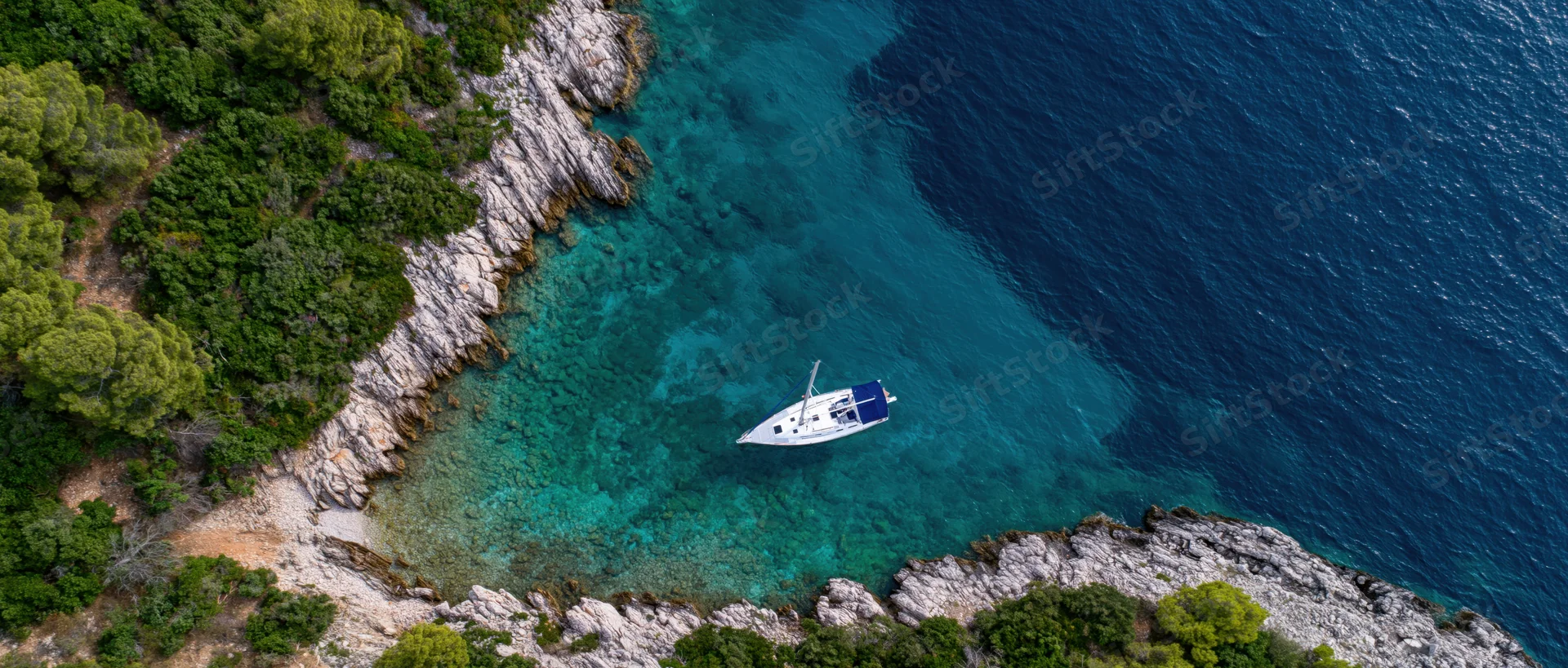 Sailboat anchored in a secluded cove surrounded by rocky cliffs and green trees, viewed from above