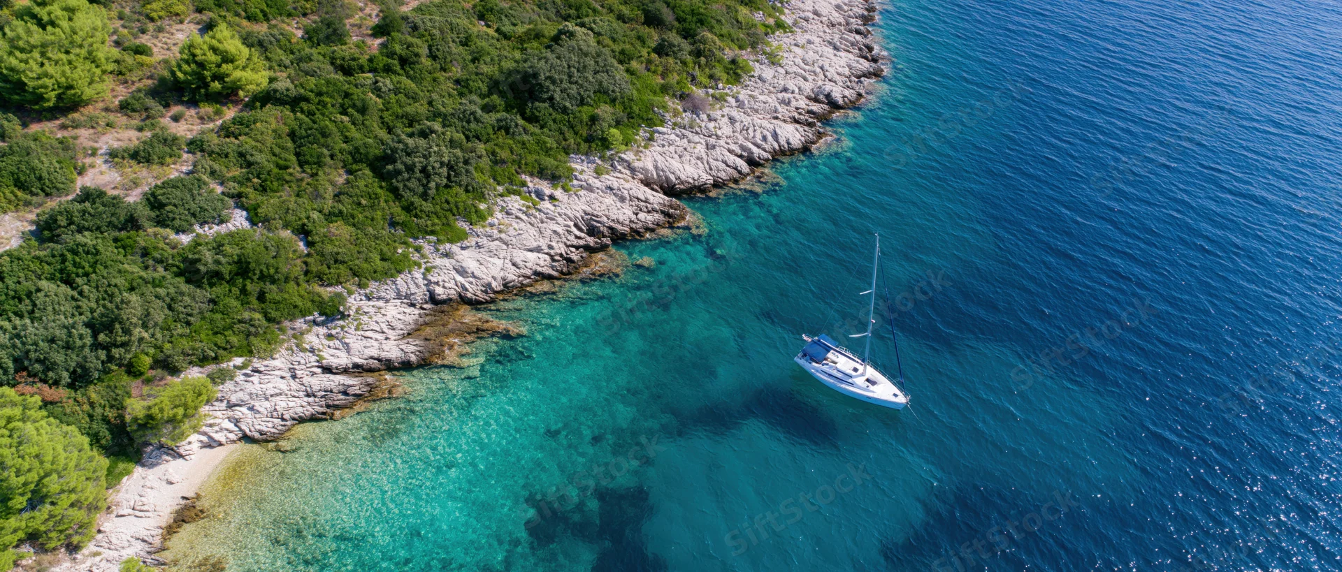 Sailboat anchored near rocky coastline with turquoise water and green vegetation from aerial view