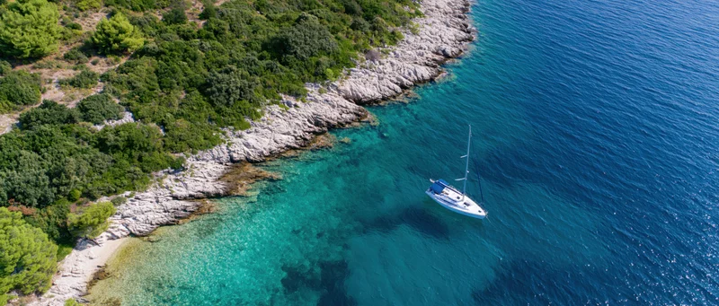 Sailboat anchored near rocky coastline with turquoise water and green vegetation from aerial view