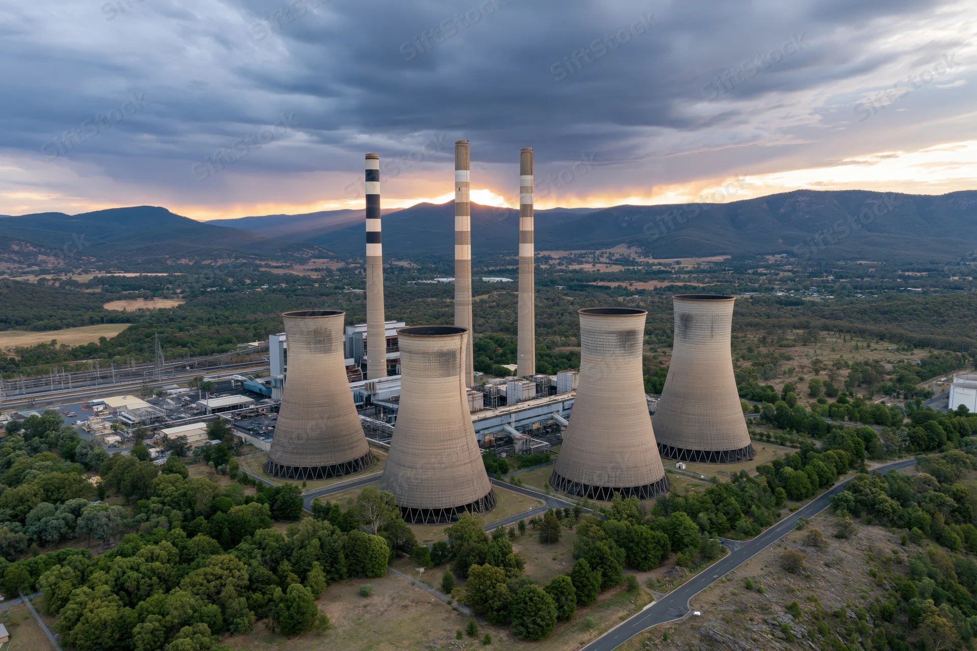 Aerial view of a coal power plant with four cooling towers and tall chimneys surrounded by mountains and greenery at sunset