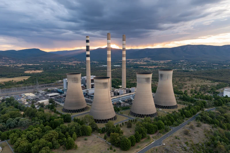 Aerial view of a coal power plant with four cooling towers and tall chimneys surrounded by mountains and greenery at sunset