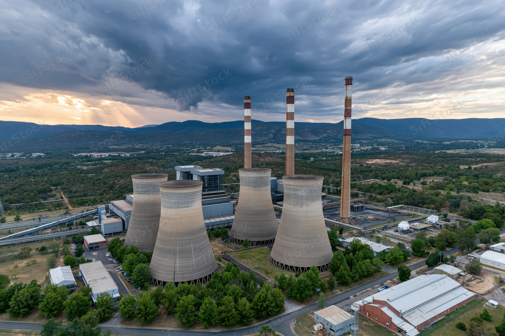 Aerial view of coal power plant with cooling towers and smokestacks surrounded by green landscape under cloudy sky at sunset