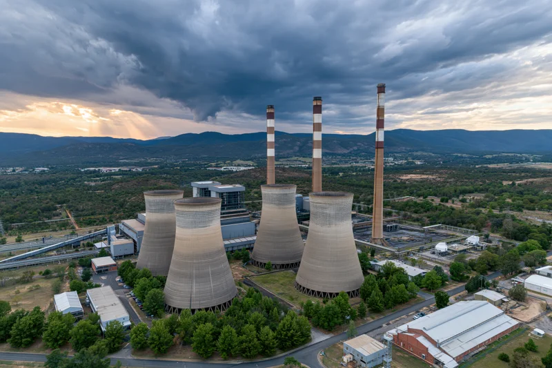 Aerial view of coal power plant with cooling towers and smokestacks surrounded by green landscape under cloudy sky at sunset