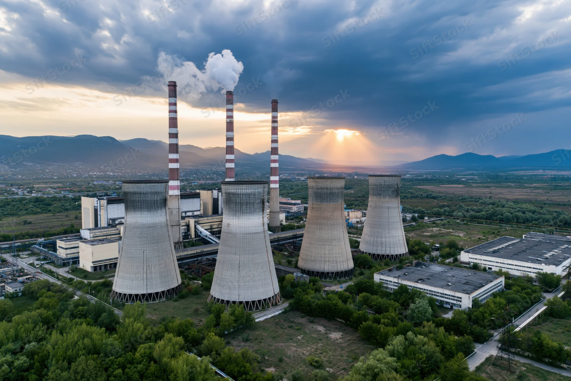 Aerial view of coal power plant with four cooling towers and smokestacks emitting steam against a sunset sky over a mountainous landscape