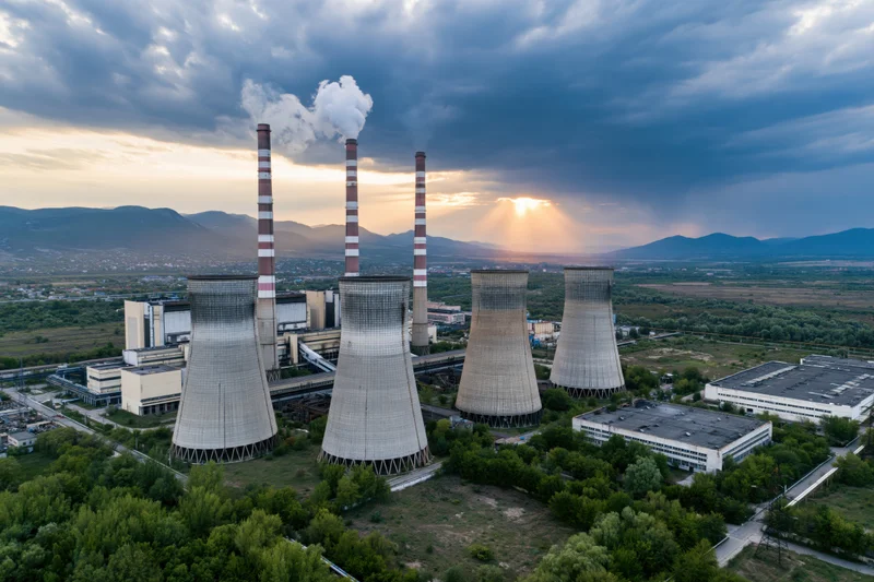 Aerial view of coal power plant with four cooling towers and smokestacks emitting steam against a sunset sky over a mountainous landscape