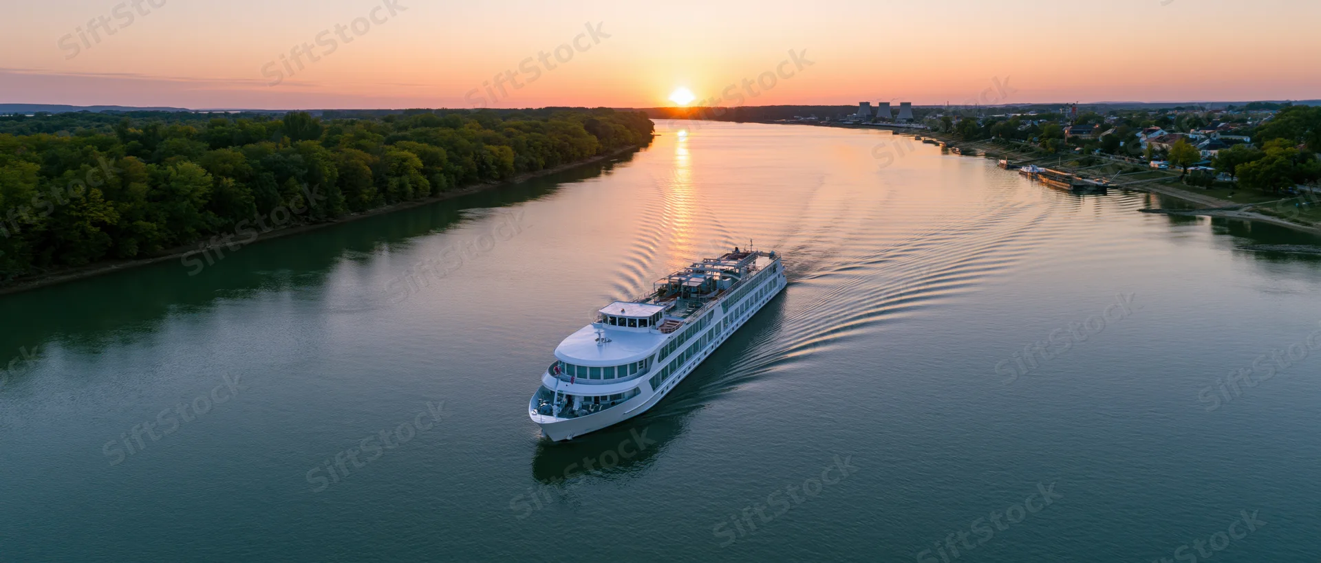 White multi-deck river cruise ship moving on calm river at sunset with trees and city along shoreline
