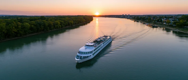 White multi-deck river cruise ship moving on calm river at sunset with trees and city along shoreline
