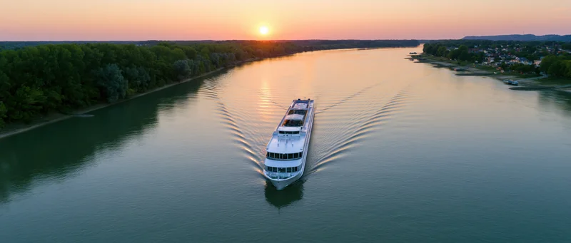 White multi-deck river cruise ship moving on calm river at sunset with trees lining the banks