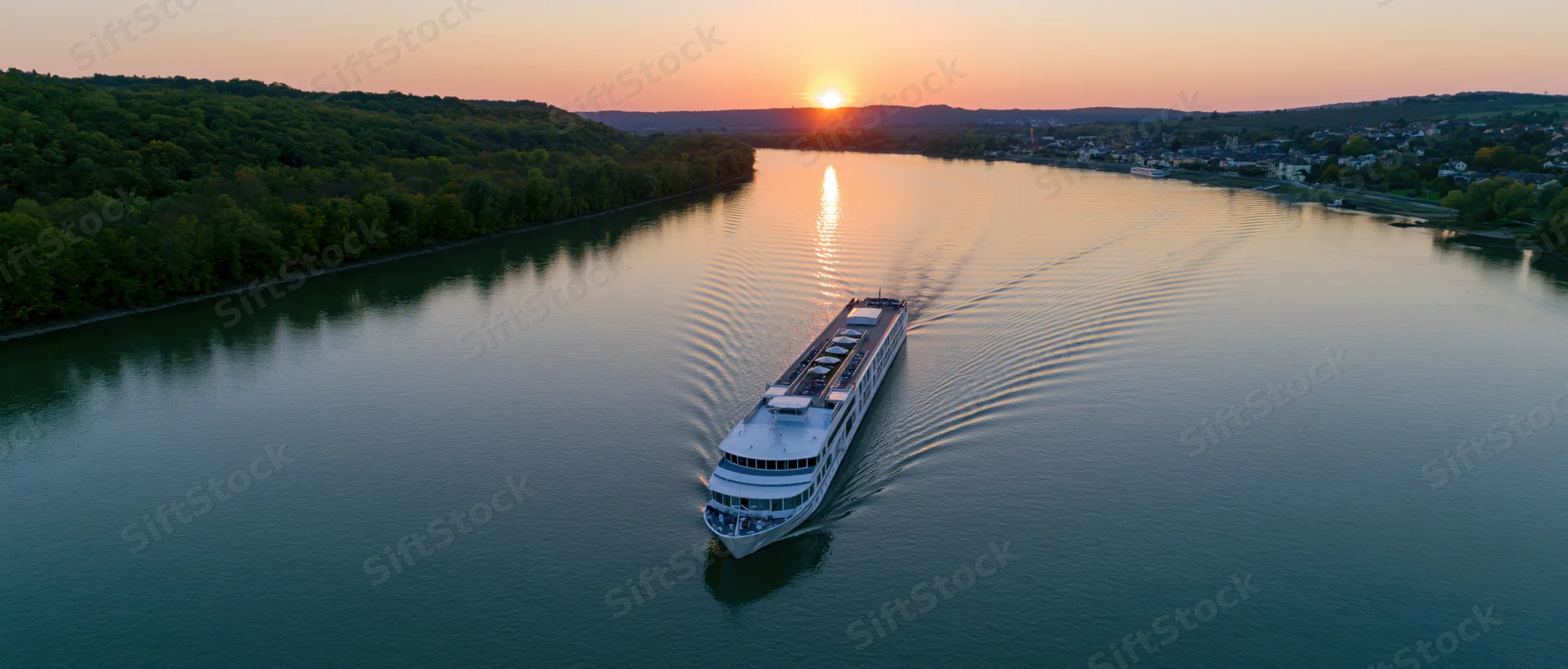 White multi-deck river cruise ship moving on calm river at sunset with forested banks and distant town