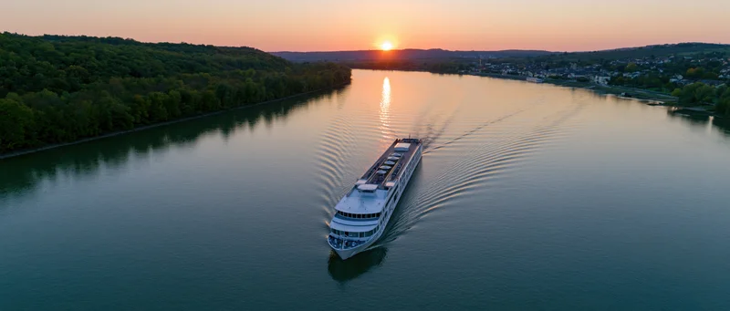 White multi-deck river cruise ship moving on calm river at sunset with forested banks and distant town