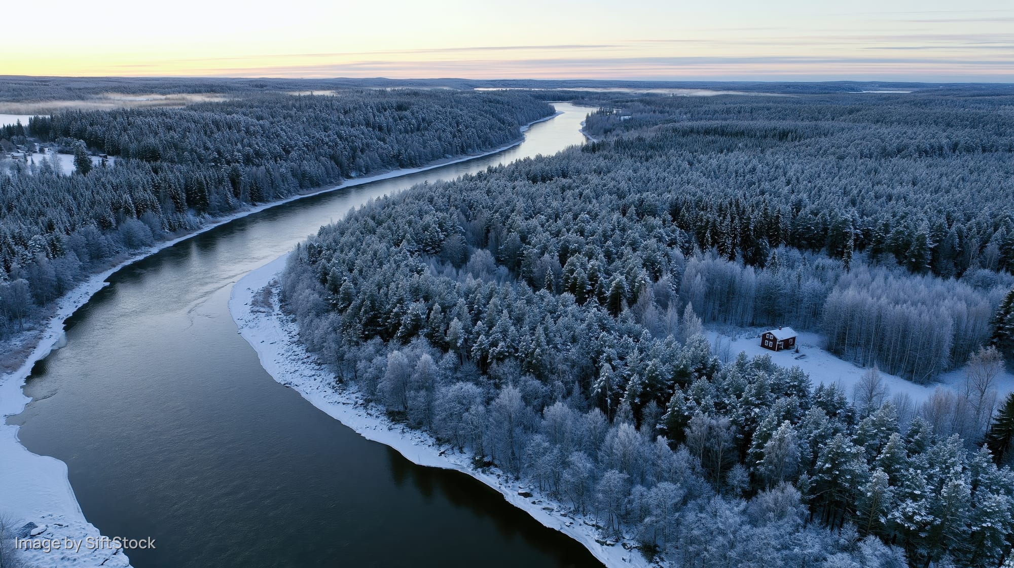 Aerial view of a winding river through snow-covered forest with a cabin in the distance at twilight