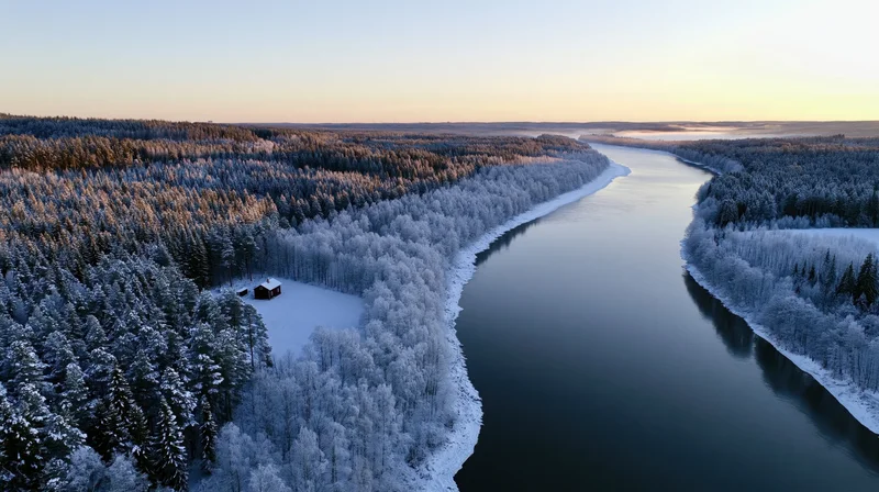 Wide river winding through snow-covered forest with cabin on bank, aerial view at sunrise