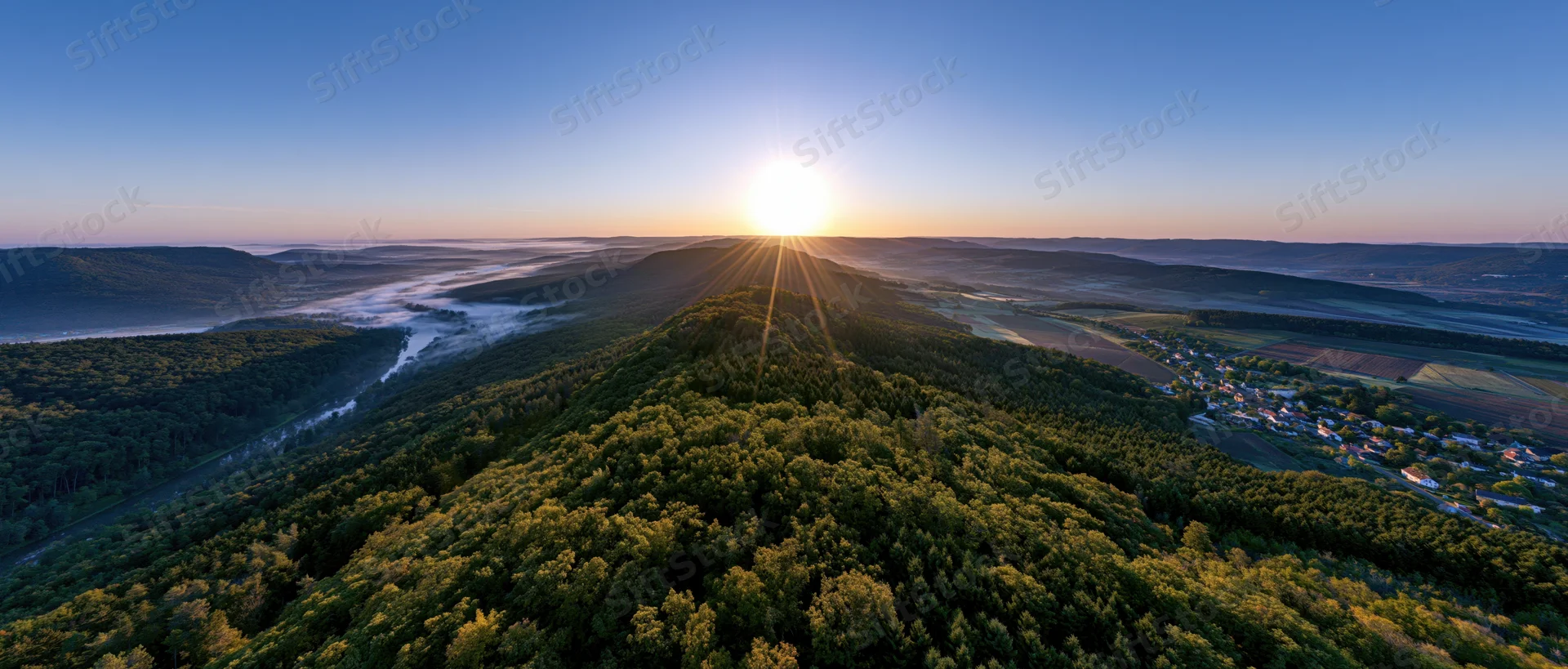 Aerial panoramic view of forested hill at sunrise with sunburst over valley and village