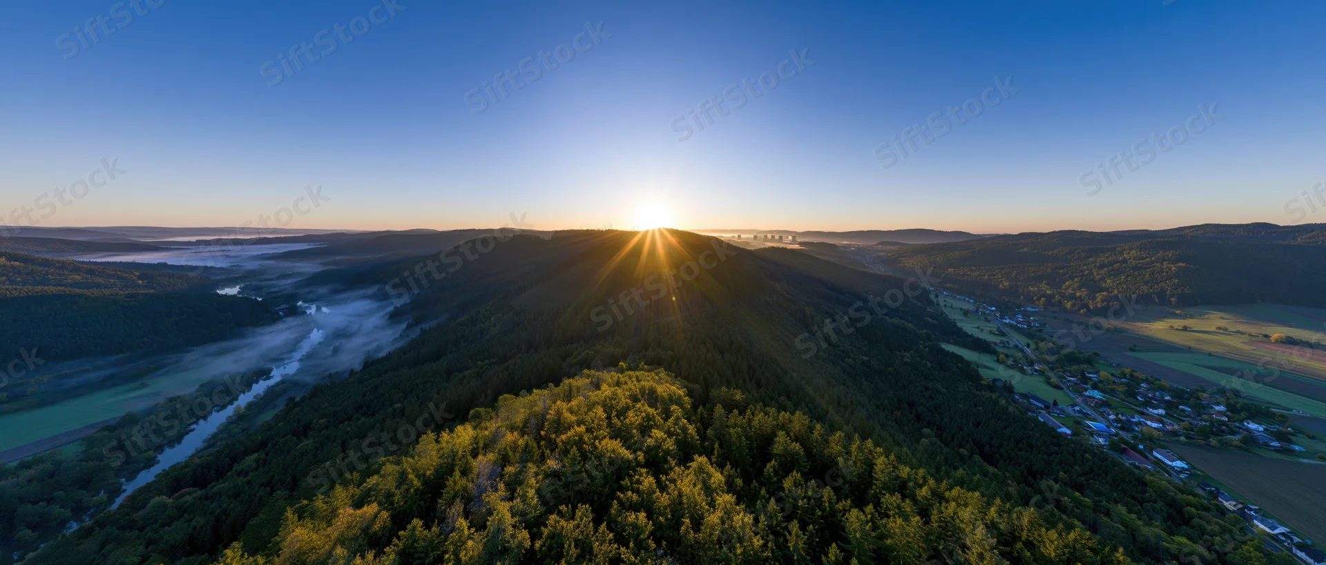 Aerial panoramic view of forested hill at sunrise with sunburst over valley and river