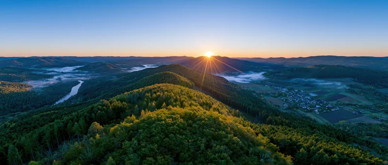 Aerial panoramic view of forested hill at sunrise with sunburst over valley and village