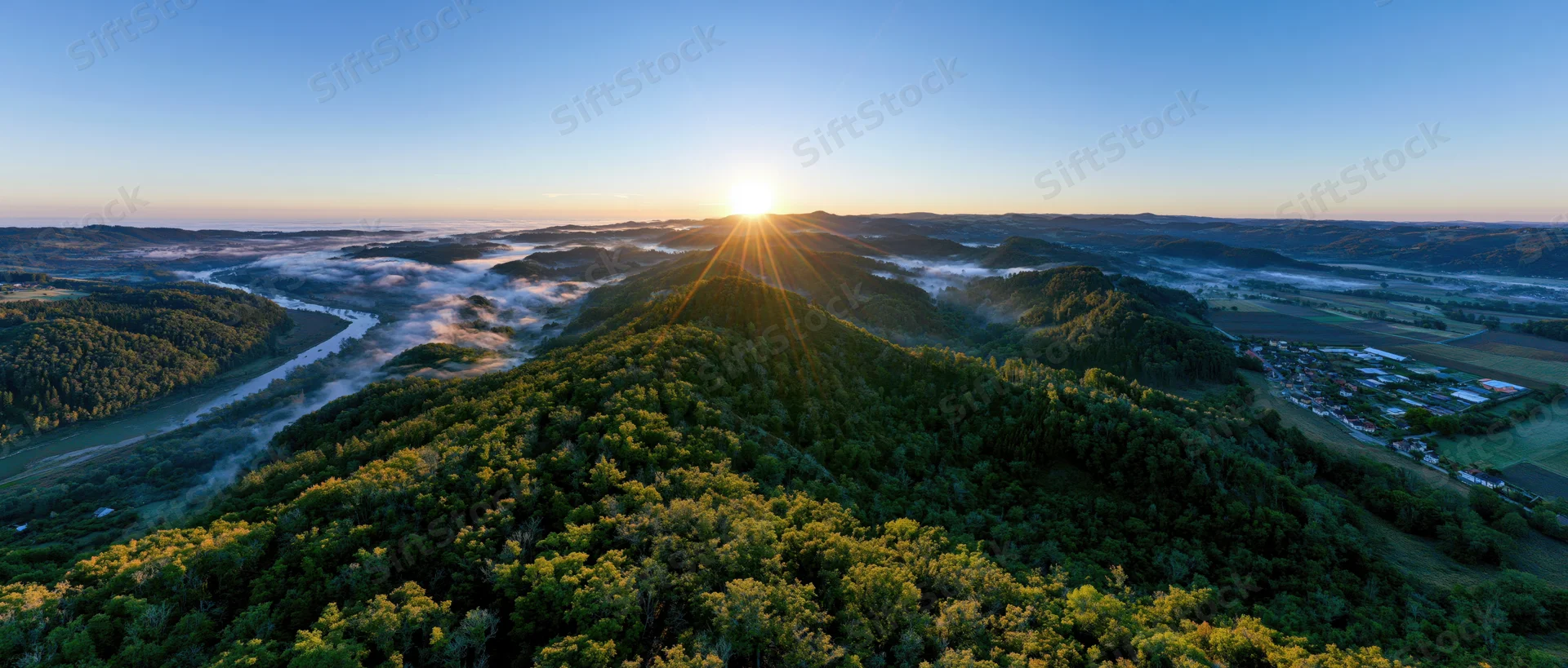 Aerial panoramic view of forested hill at sunrise with sunburst over valley and river