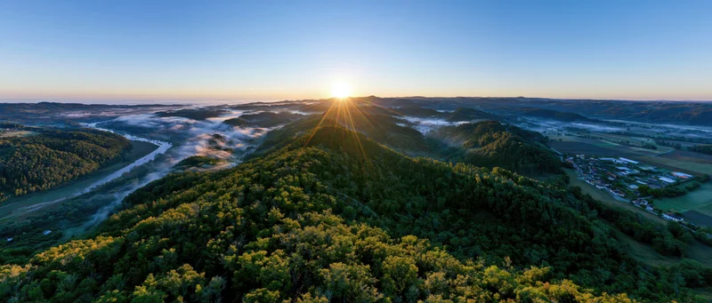 Aerial panoramic view of forested hill at sunrise with sunburst over valley and river