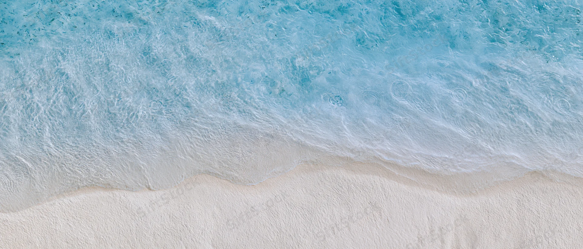 Aerial top-down view of a wave breaking onto pale cream sand at a shallow shoreline