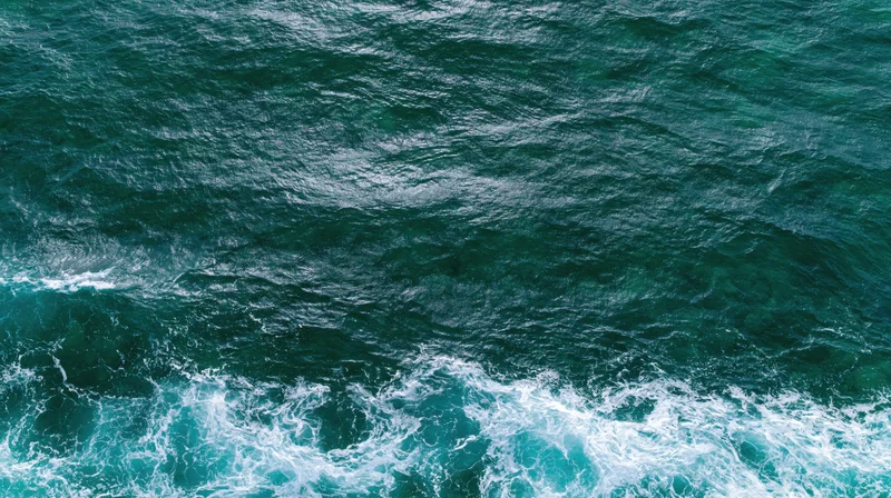 Aerial top-down view of ocean surface with rippling waves and white foam along the edge