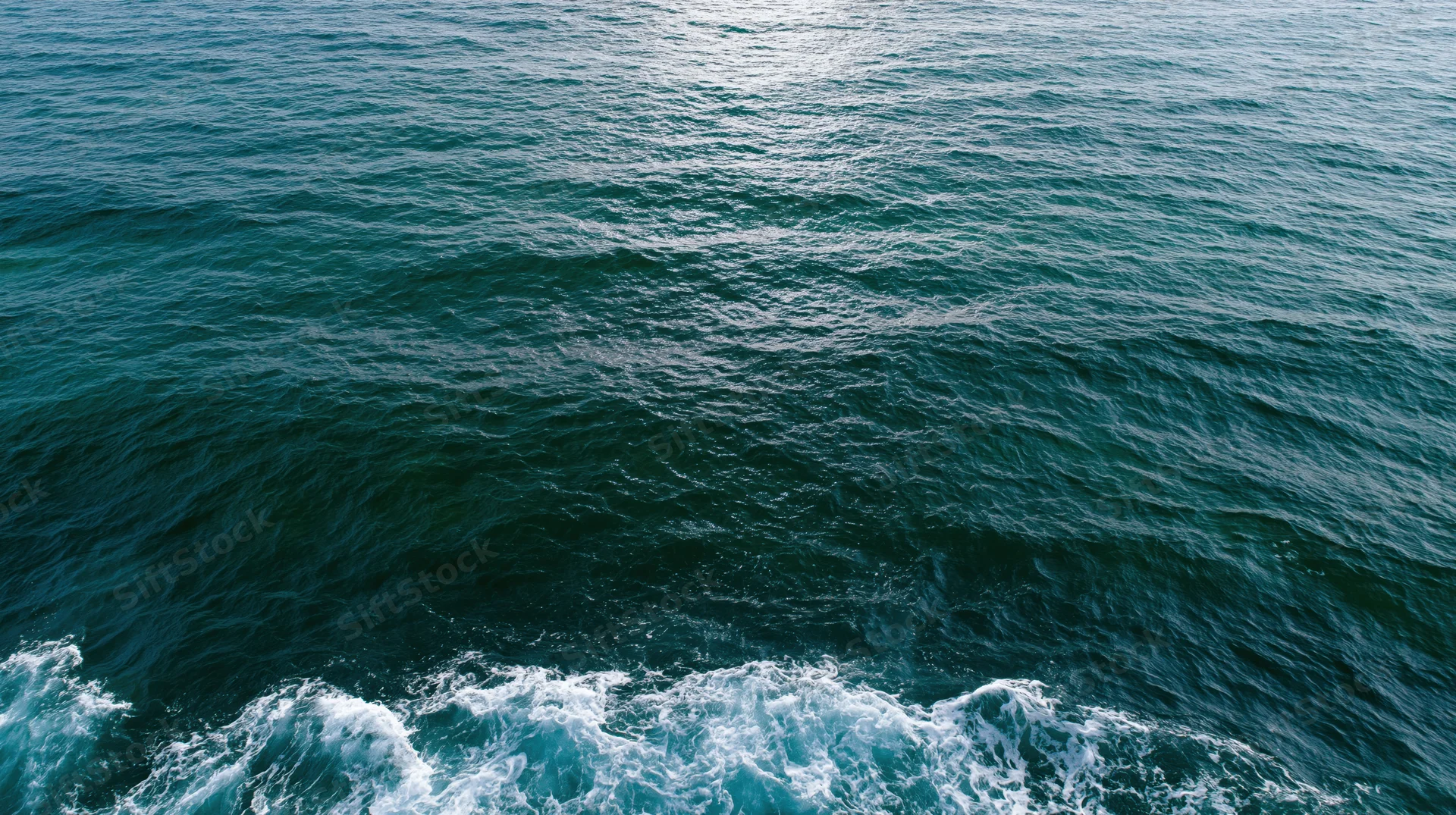 Aerial top-down view of open ocean surface with small ripples and white foam along the bottom edge