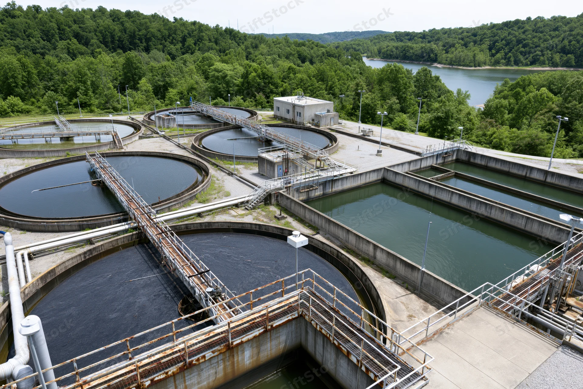 Aerial view of wastewater treatment facility with circular and rectangular tanks surrounded by trees and a river