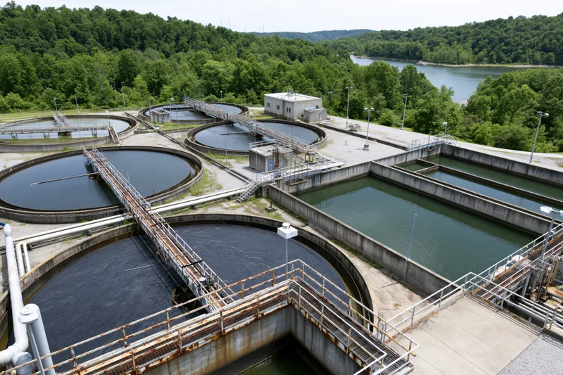Aerial view of wastewater treatment facility with circular and rectangular tanks surrounded by trees and a river