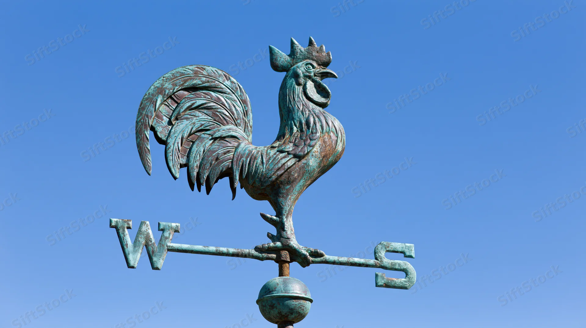 Aged copper rooster weather vane with cardinal direction arrows against blue sky