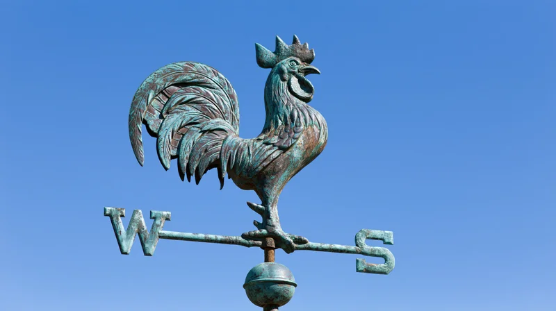 Aged copper rooster weather vane with cardinal direction arrows against blue sky