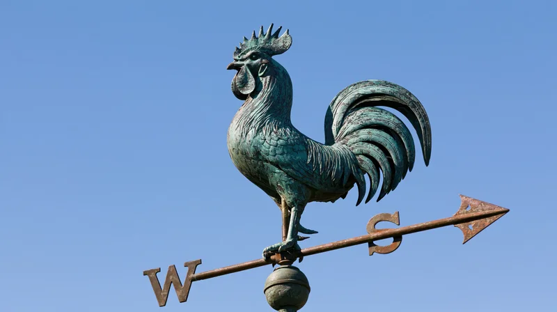 Aged copper rooster weather vane with cardinal direction arrow against blue sky