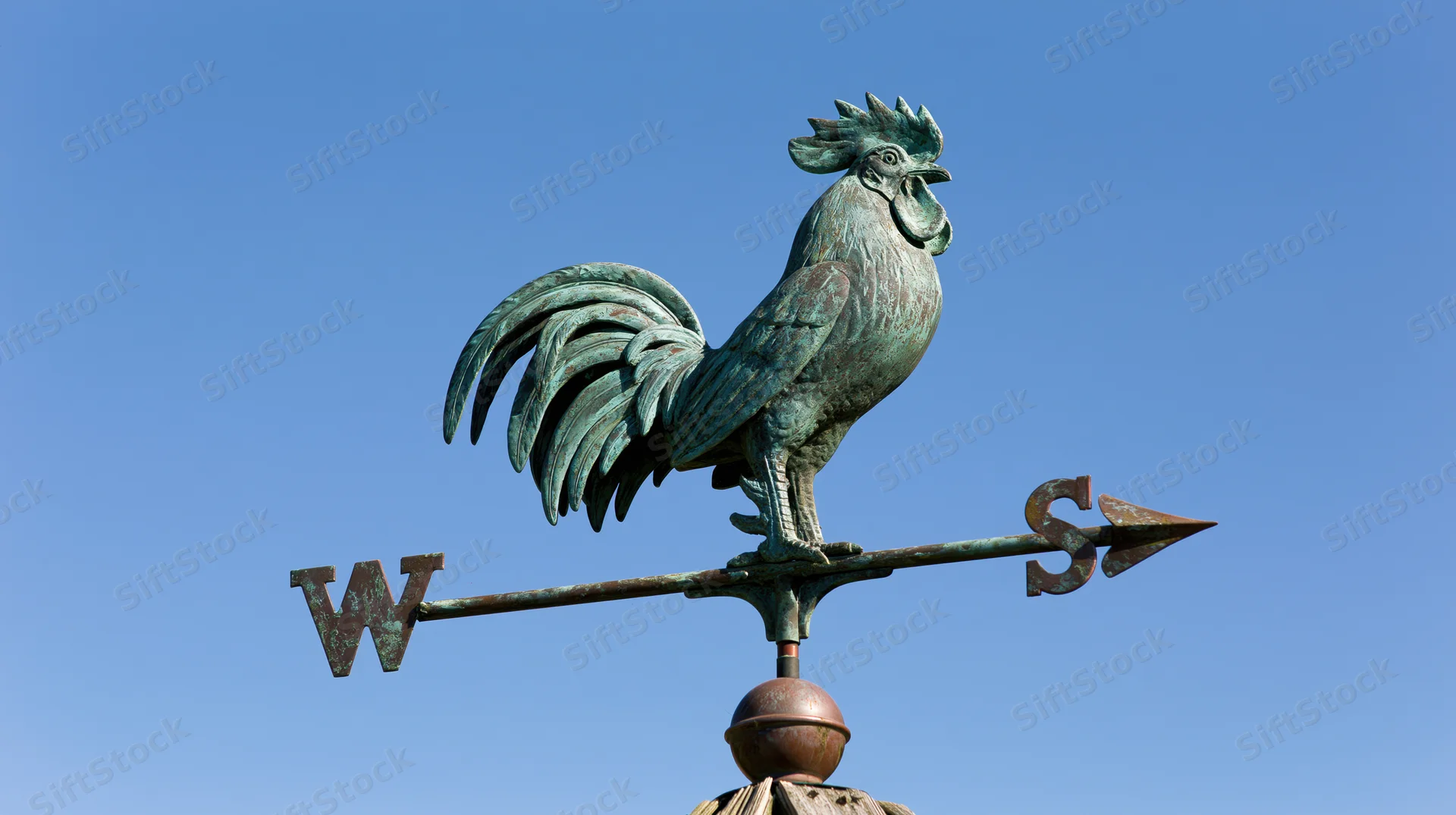 Aged copper rooster weather vane with cardinal direction arrows against blue sky