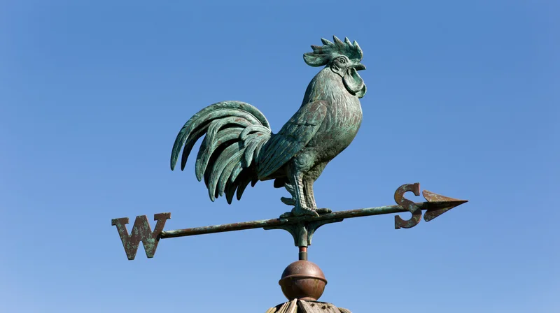 Aged copper rooster weather vane with cardinal direction arrows against blue sky