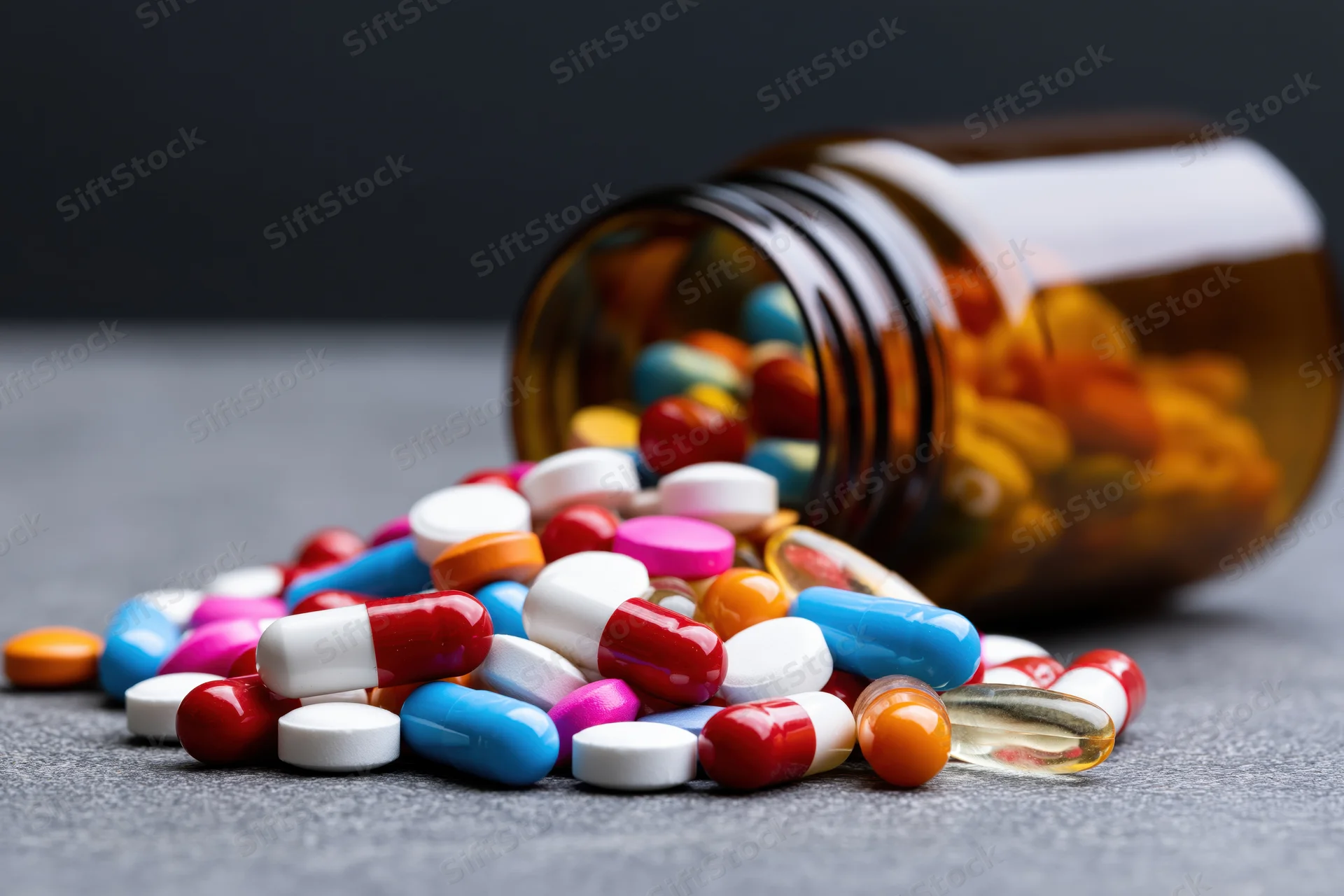 Spilled colorful pills and capsules from an amber prescription bottle on a gray surface